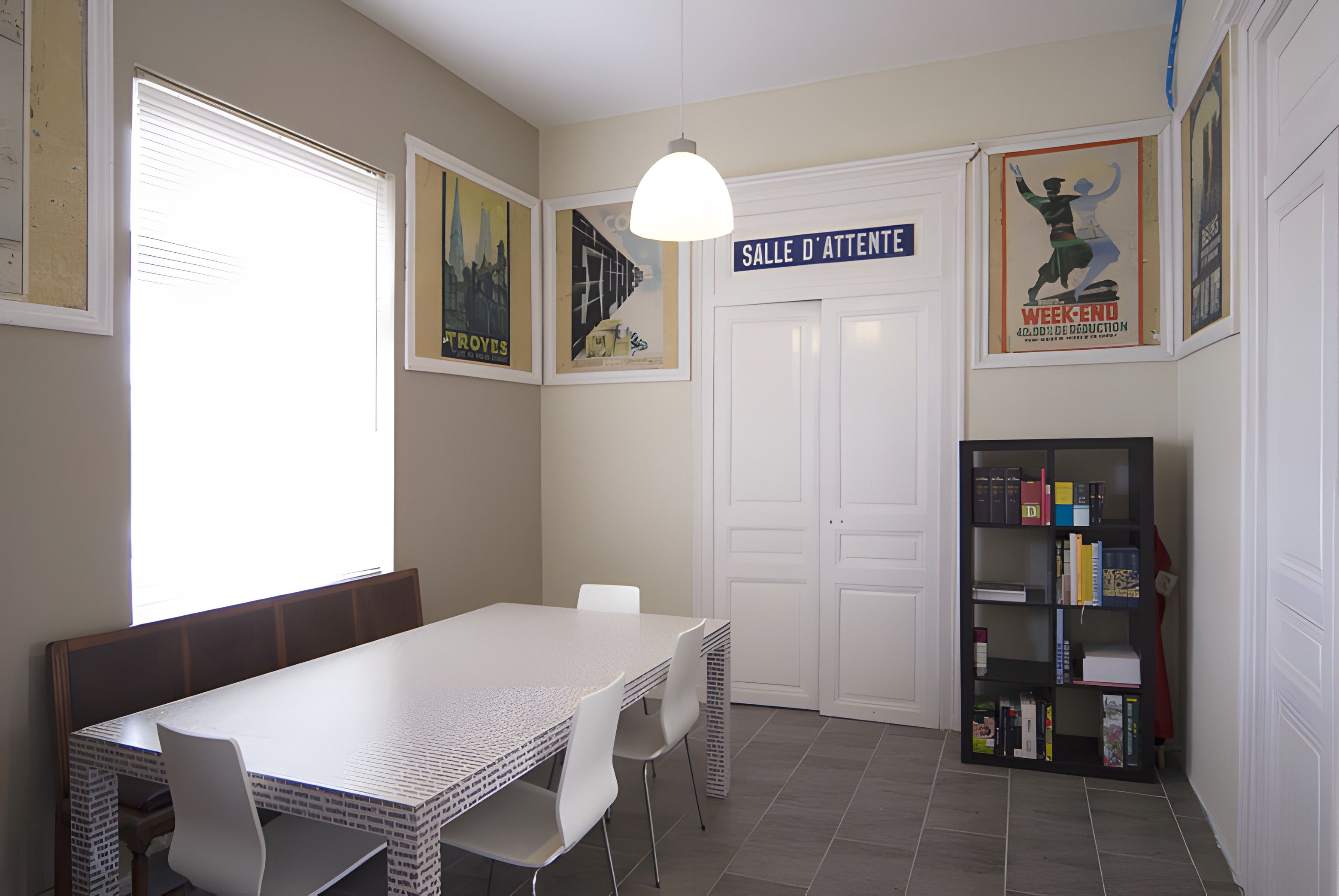 Un coin repas avec une table et des chaises blanches, une bibliothèque et une porte portant l’inscription « SALLE D’ATTENTE ».