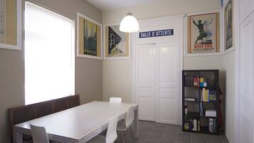Un coin repas avec une table et des chaises blanches, une bibliothèque et une porte portant l’inscription « SALLE D’ATTENTE ».
