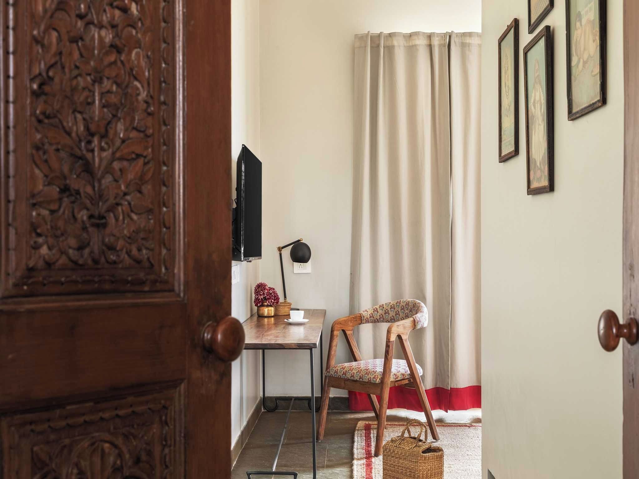 Um quarto com porta de madeira, uma escrivaninha com cadeira, televisão e cortina.