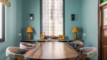 Uma sala de jantar com uma mesa de madeira redonda, rodeada por cadeiras com padrão floral, uma janela com cortinas estampadas e duas luminárias fixadas na parede.