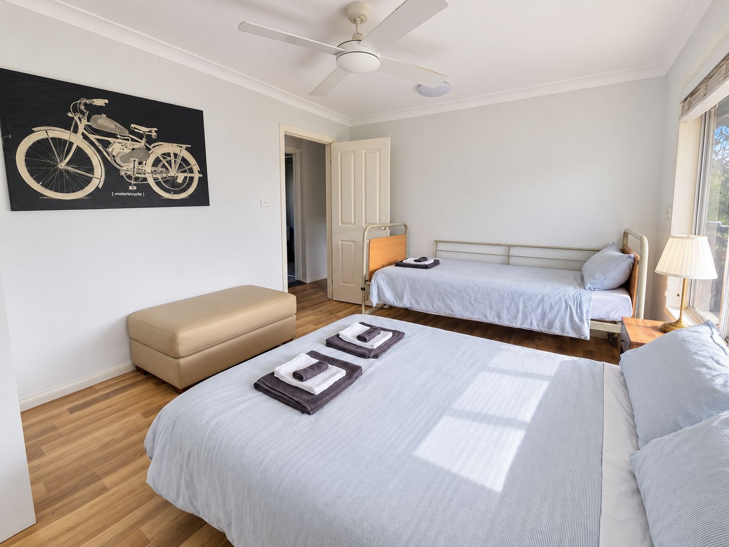 Quarto com duas camas, ventoinha no teto, janela com vista e uma obra de arte na parede retratando uma motocicleta.