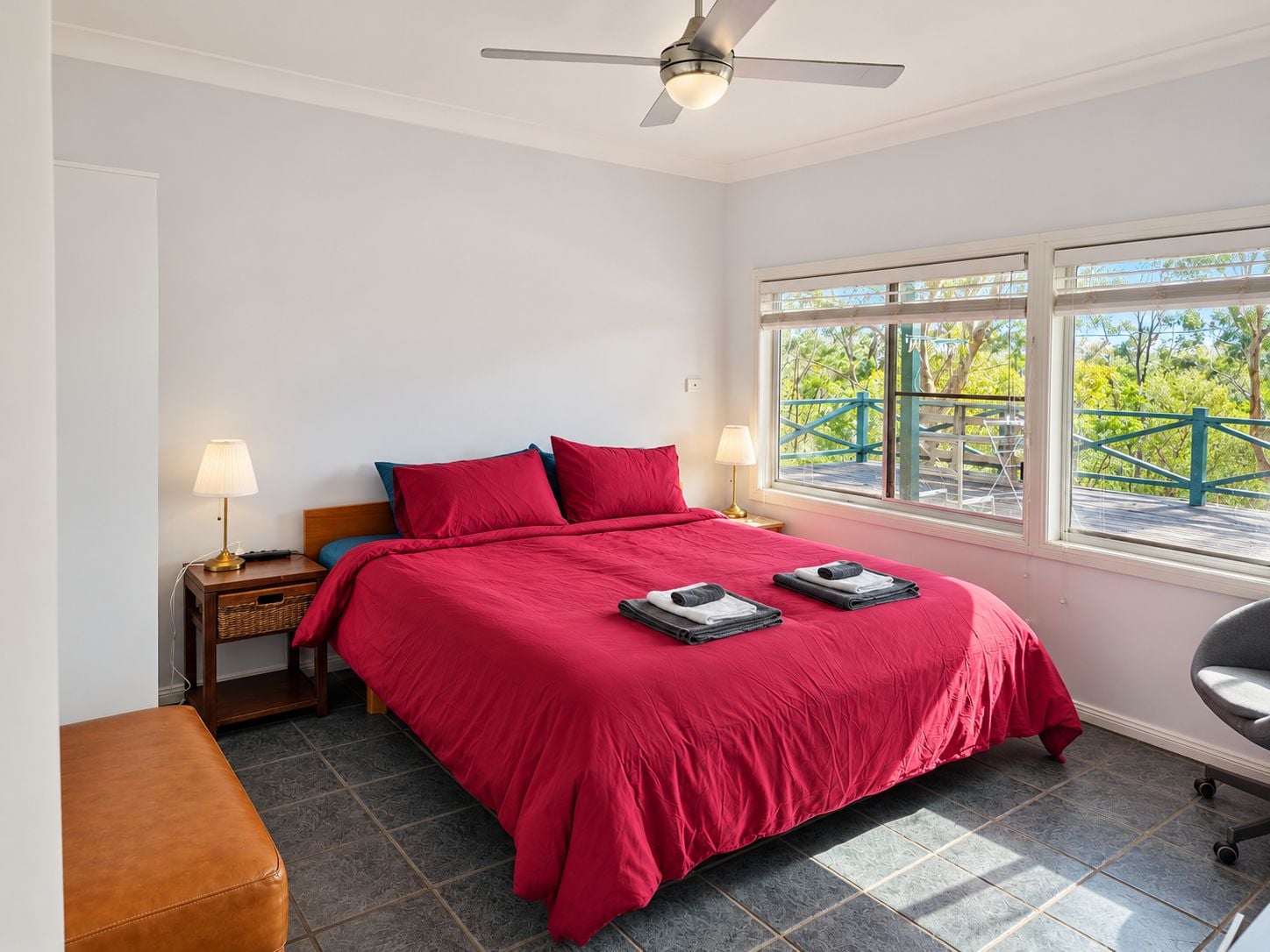 A bedroom with a bed, two bedside lamps, a nightstand, a bench, and a window with a view of greenery.
