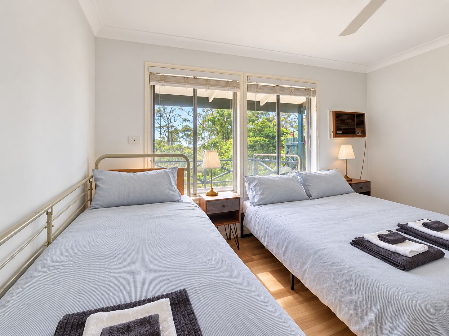 Une chambre double avec deux lits, une fenêtre avec des stores, une table de chevet avec une lampe, et une vue sur la verdure.