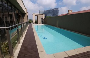 Una piscina con cubierta de madera, barandillas de vidrio y vistas a edificios modernos al fondo.