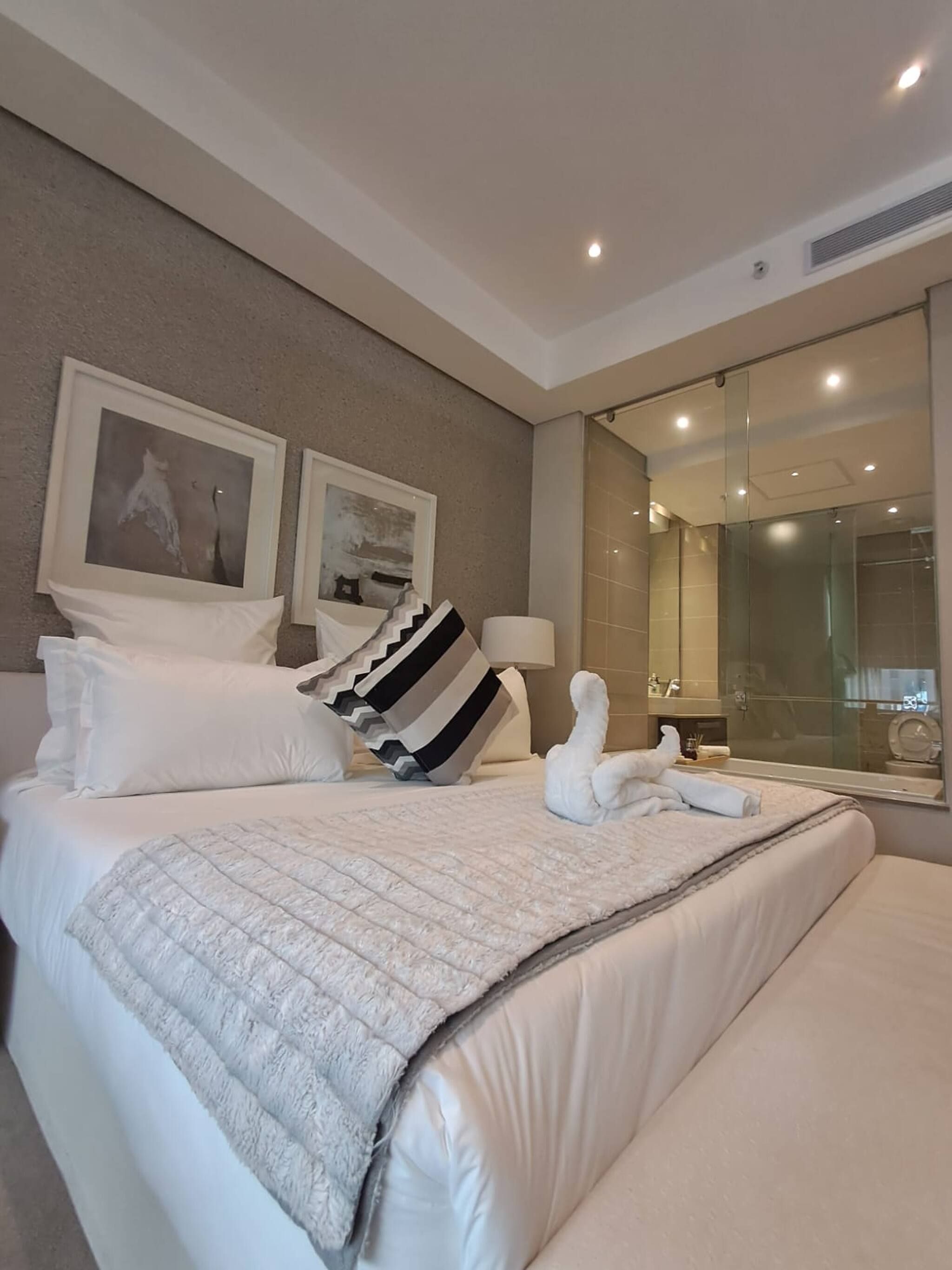 Habitación de hotel con una cama grande, dos cuadros en la pared, cabina de ducha de vidrio y un espejo que refleja la habitación.