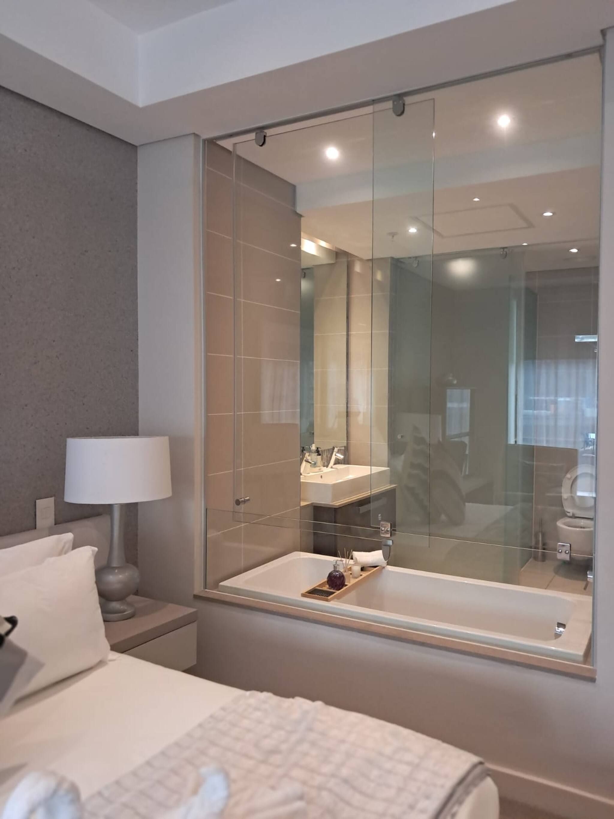 Una habitación de hotel moderna con una cama grande, una mesita de noche con una lámpara, un baño con paredes de vidrio y una bañera, y un espejo grande.