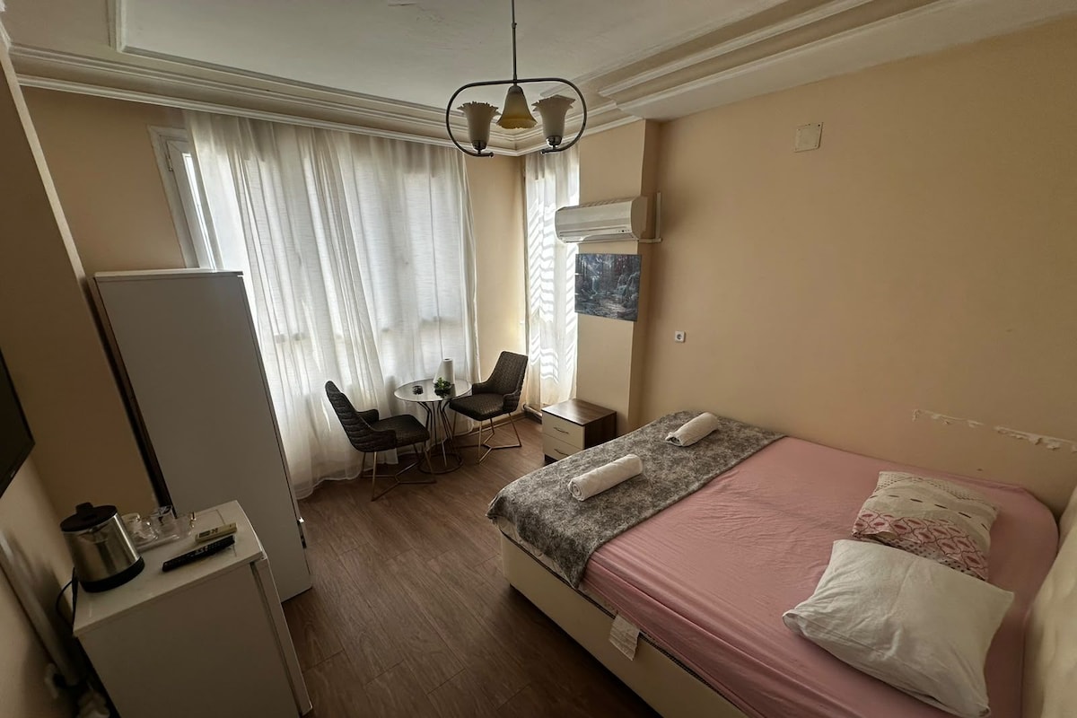 Quarto de hotel com cama, escrivaninha, cadeira, mesa pequena e janela com cortinas.