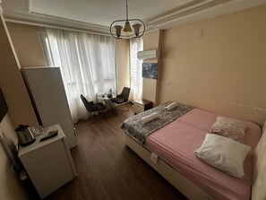 Quarto de hotel com cama, escrivaninha, cadeira, mesa pequena e janela com cortinas.