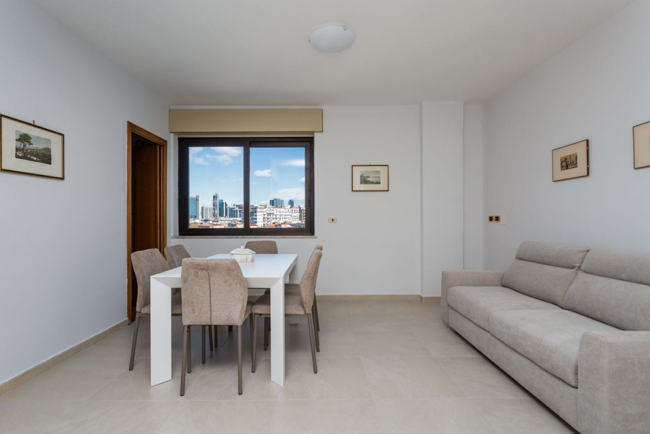 A modern living room with a dining area, a sofa, and a view of the cityscape through a large window.