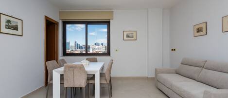 A modern living room with a dining area, a sofa, and a view of the cityscape through a large window.