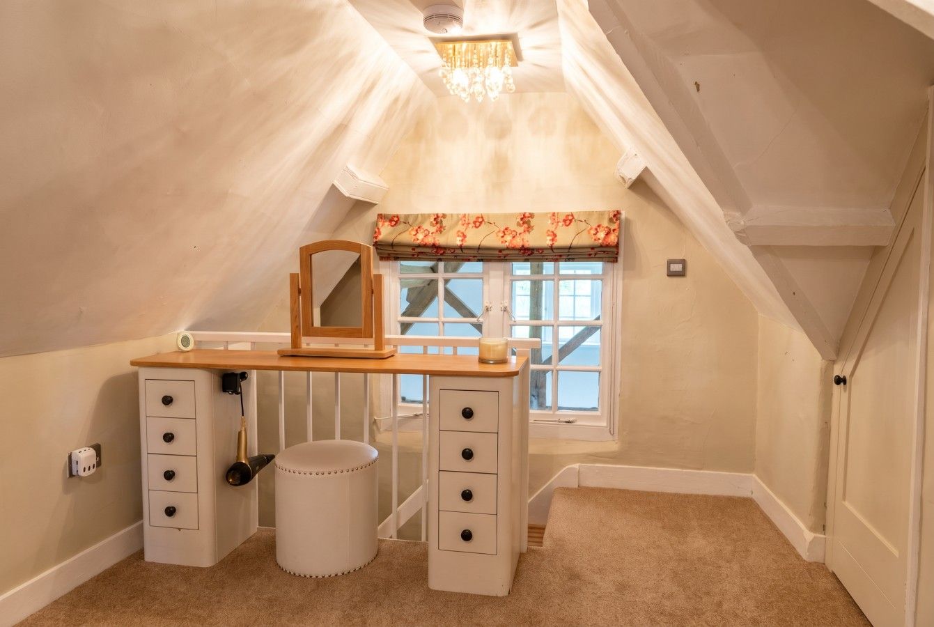 A bedroom with a sloped ceiling, a desk with drawers, a chair, a small round ottoman, a window with a floral valance, and a door.