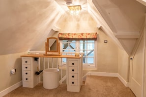 Ein Schlafzimmer mit schrägem Dach, einem Schreibtisch mit Schubladen, einem Stuhl, einem kleinen runden Hocker, einem Fenster mit Blumenrollo und einer Tür.