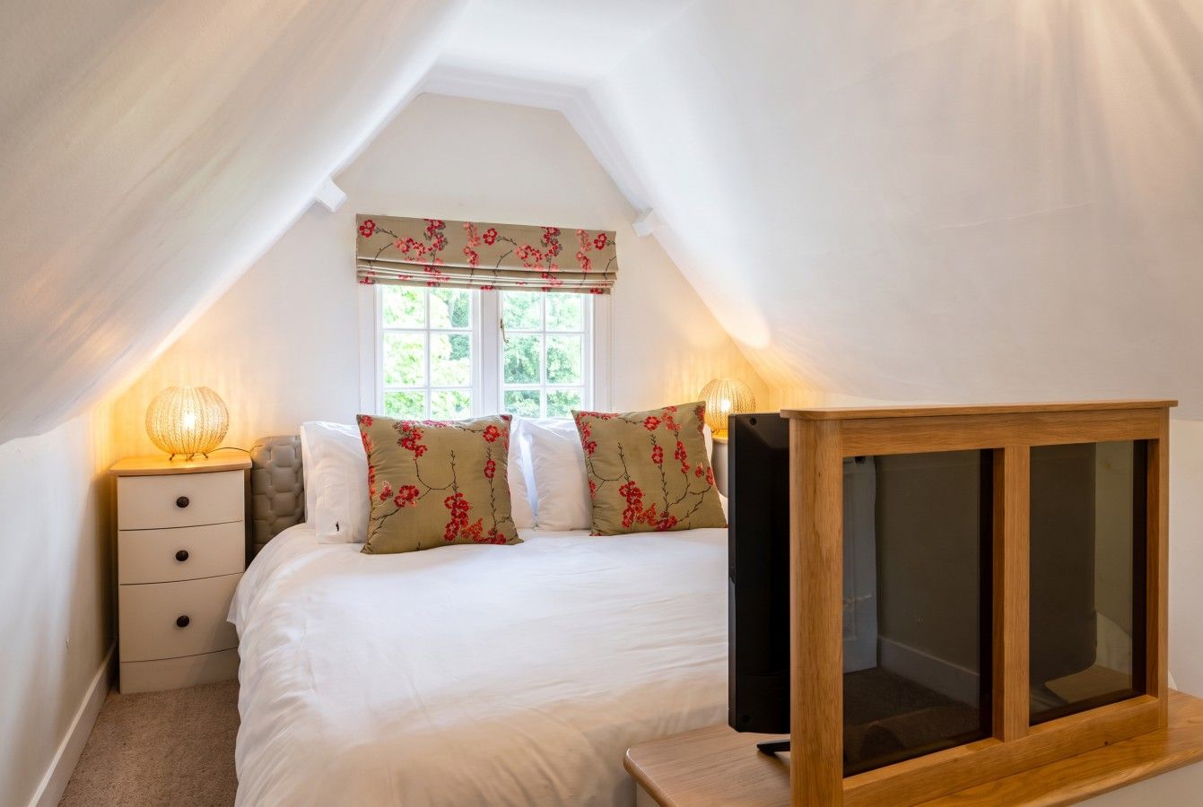 Ein Schlafzimmer mit einem Bett, einem Nachttisch, einem Fernseher und einem Fenster mit floralem Vorhang.