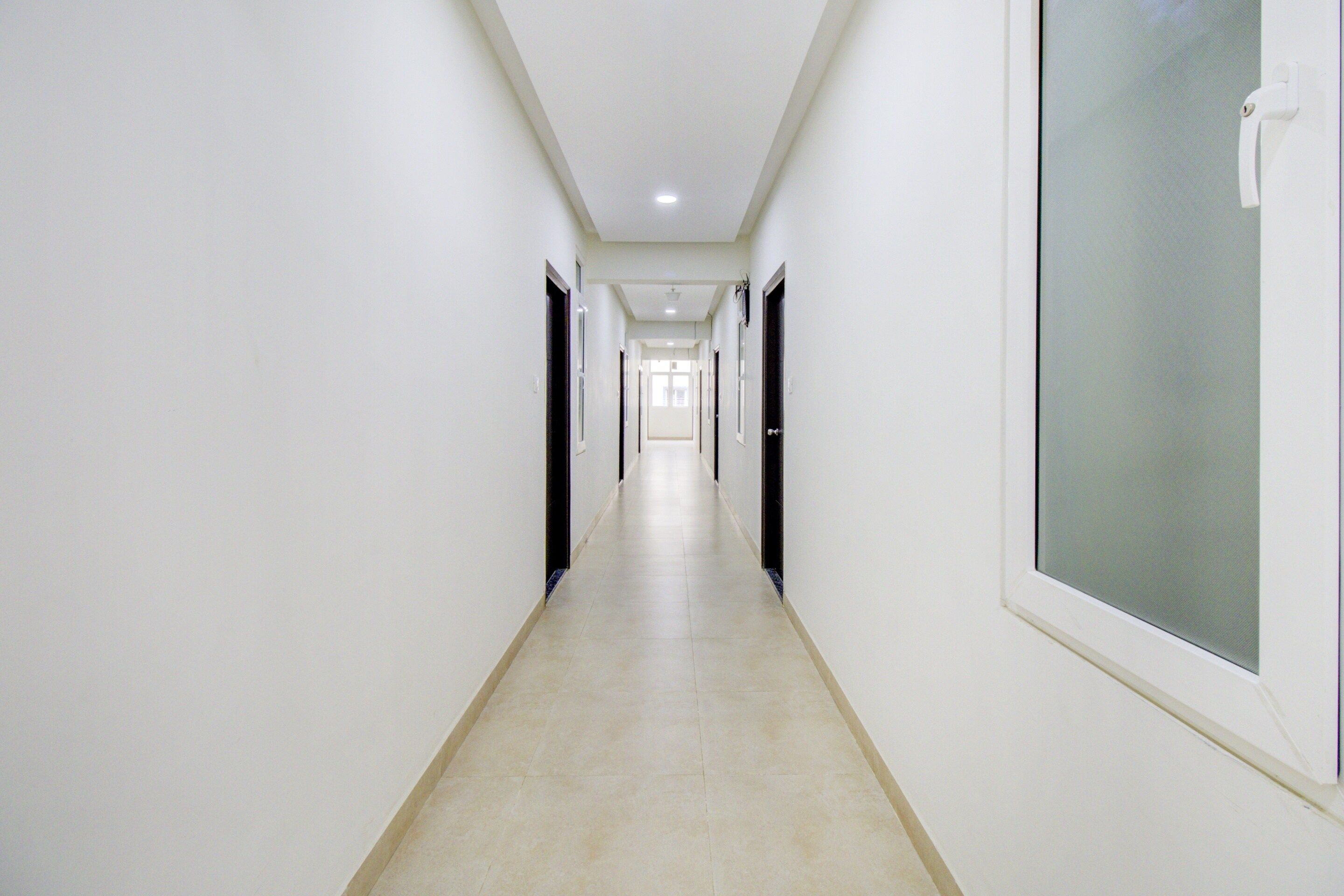 Un pasillo largo y estrecho con paredes y puertas blancas a ambos lados, que conduce a una habitación luminosa al final.