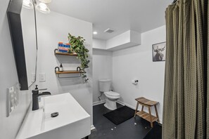 Et moderne badeværelse med en hvid vask, toilet og en lille træskammel. Der er en hylde med en plante og en æske mærket 'FIRST AID'.