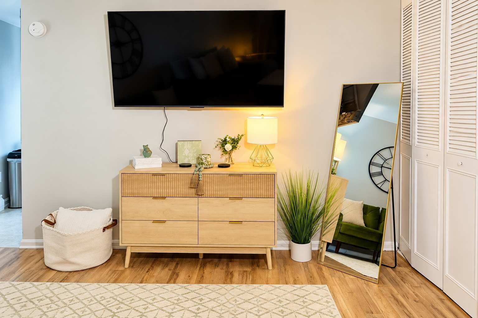 A modern living room with a flat-screen TV mounted on the wall, a wooden cabinet with drawers, a lamp, a potted plant, and a mirror.
