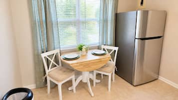 A compact kitchen with a wooden table, two chairs, a stainless steel refrigerator, and a window with curtains.