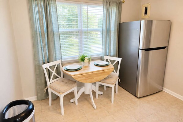 A compact kitchen with a wooden table, two chairs, a stainless steel refrigerator, and a window with curtains. - Exclusive Queen Suite near Downtown Louisville! (Clarksville)