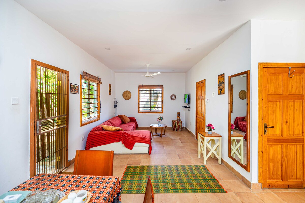 A room with a bed, a table, and a chair. There is a window with a view of greenery outside.