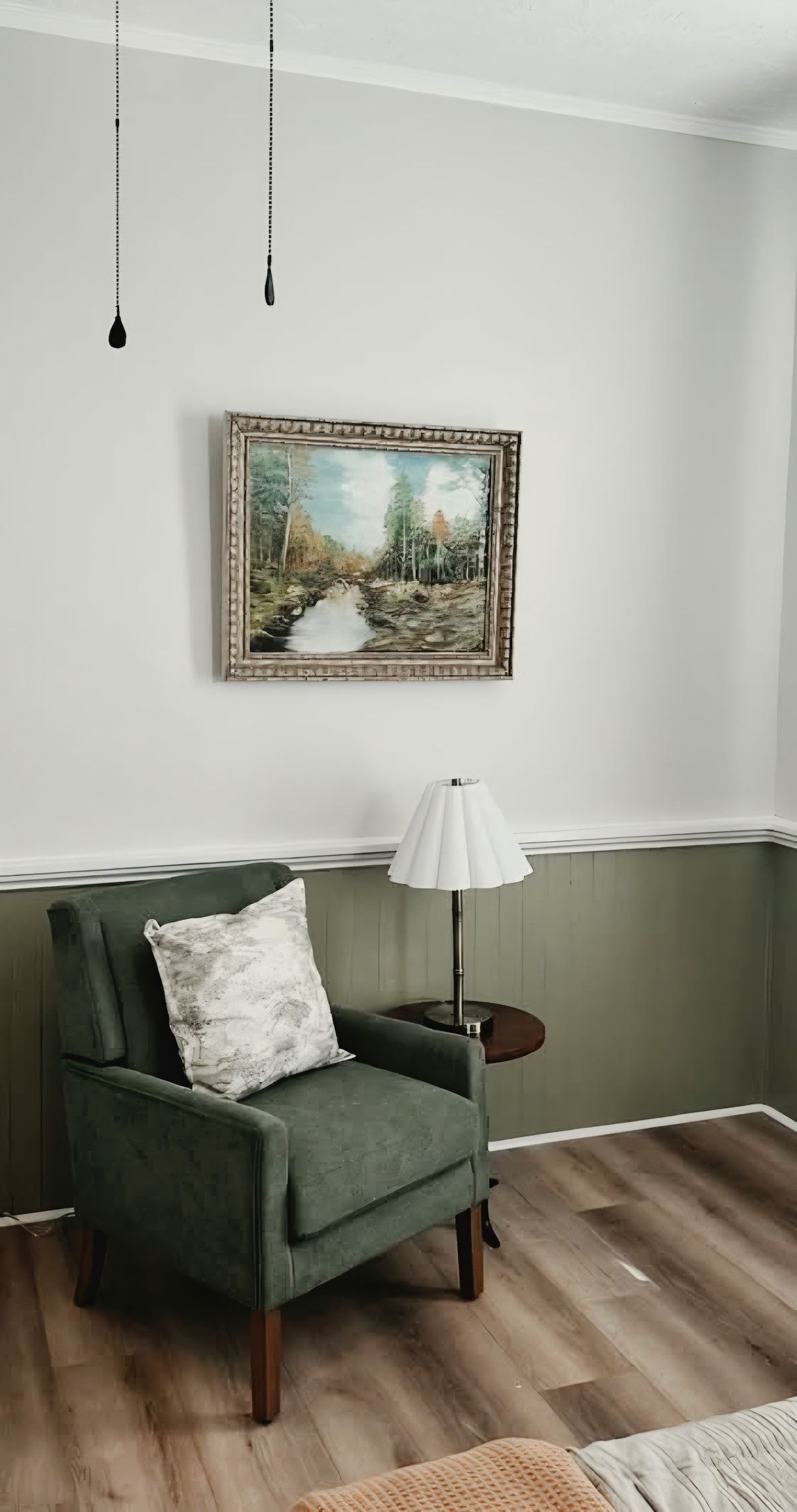 Una habitación con un sillón verde, una lámpara blanca y un cuadro de paisaje en la pared.