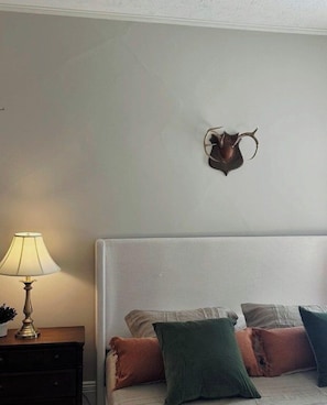 Un dormitorio con cama, almohadas, una mesita de noche con lámpara y una cabeza de animal montada en la pared.