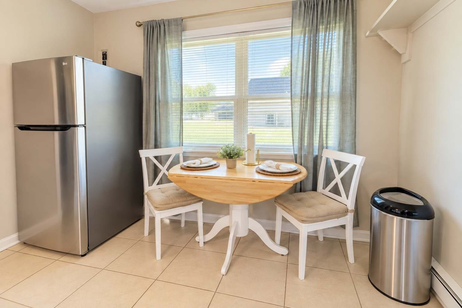 A kitchen with a stainless steel refrigerator, a wooden dining table with two chairs, and a trash bin.