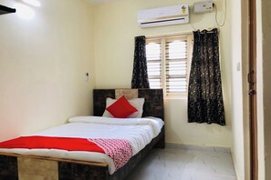 Uma cama de solteiro com cabeceira de madeira, travesseiro vermelho e cobertor branco. Há uma janela com cortinas pretas e um ar-condicionado acima.