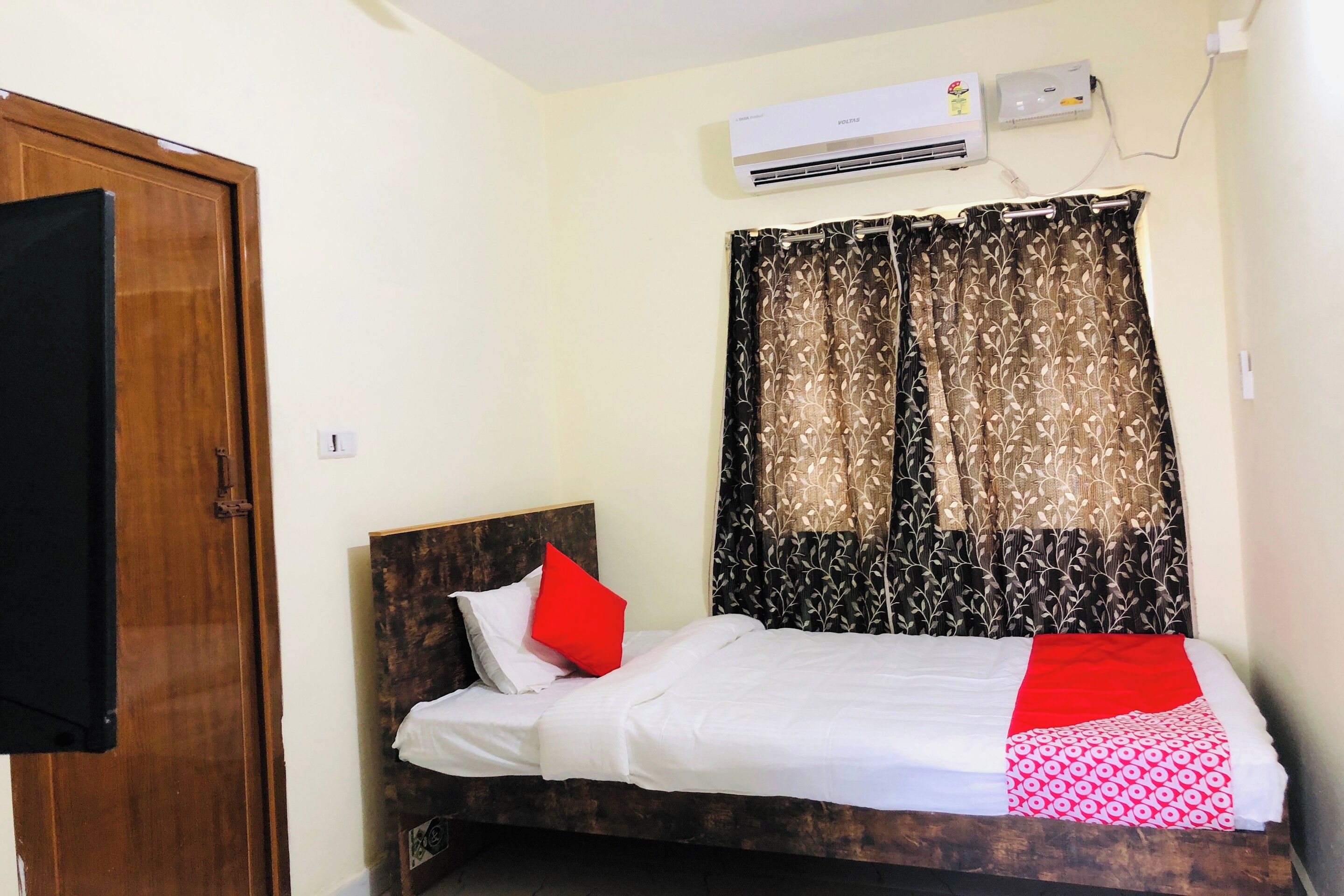 A single bed with a wooden headboard, white bedding, a red pillow, and a patterned red and white throw. An air conditioning unit is mounted on the wall above the bed.