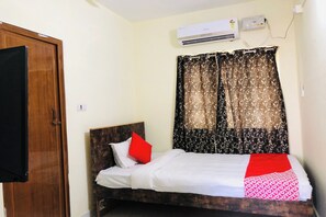 Cama de solteiro com cabeceira de madeira, roupa de cama branca, travesseiro vermelho e manta estampada em vermelho e branco. Um aparelho de ar condicionado está fixado na parede acima da cama.