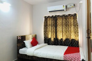 Uma cama de solteiro com cabeceira, um travesseiro vermelho e um cobertor branco. Há uma janela com cortina e um aparelho de ar condicionado fixado na parede.
