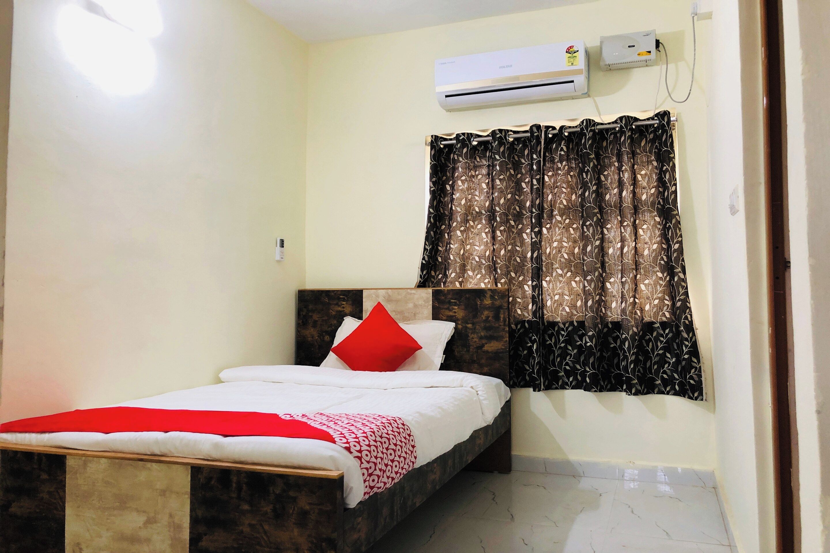 A bedroom with a bed, a window with patterned curtains, an air conditioning unit, and a wall-mounted light.