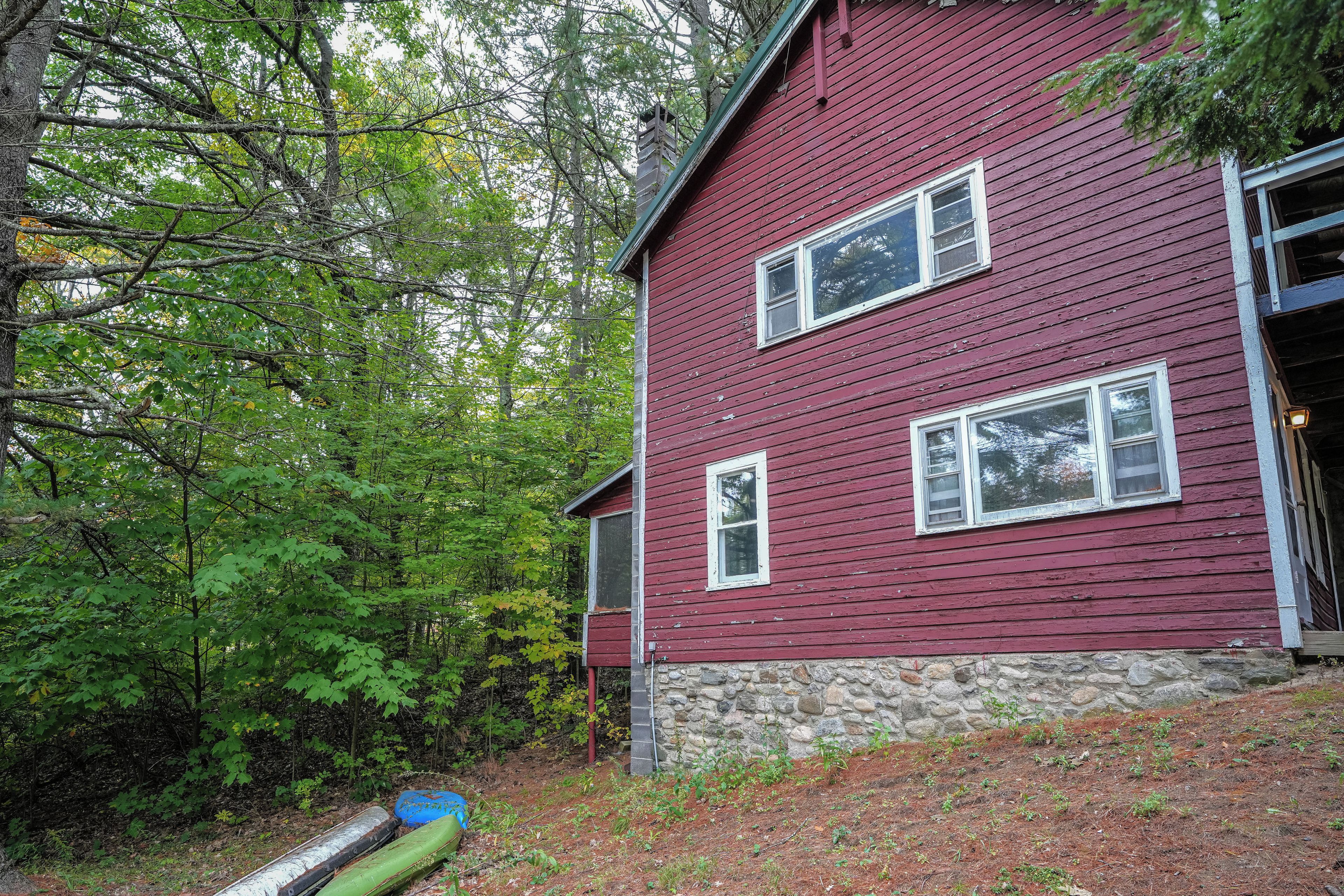 A two-story red wooden house with a stone foundation, surrounded by dense green foliage.