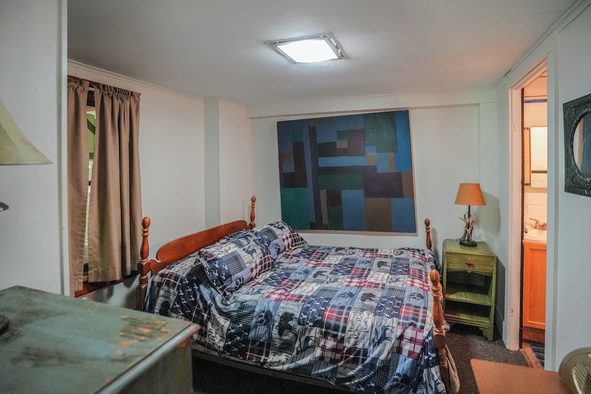 A bedroom with a bed, a nightstand, a lamp, a painting, and a door leading to another room.