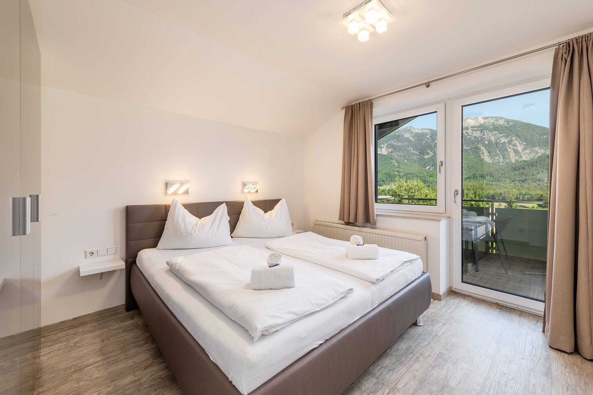 A modern bedroom with a large bed, a view of mountains through the window, and a balcony.