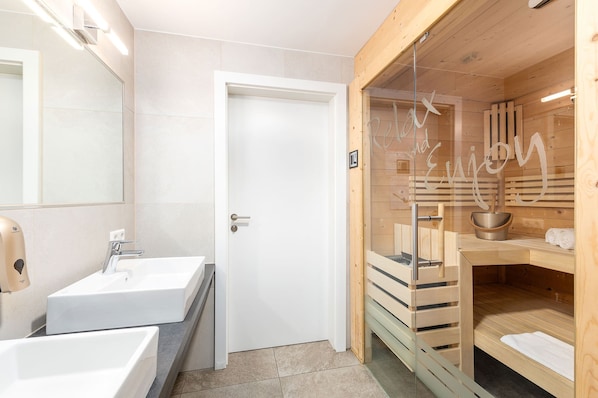 Un baño moderno con un lavamanos blanco, un espejo y una zona de sauna con paredes de vidrio.