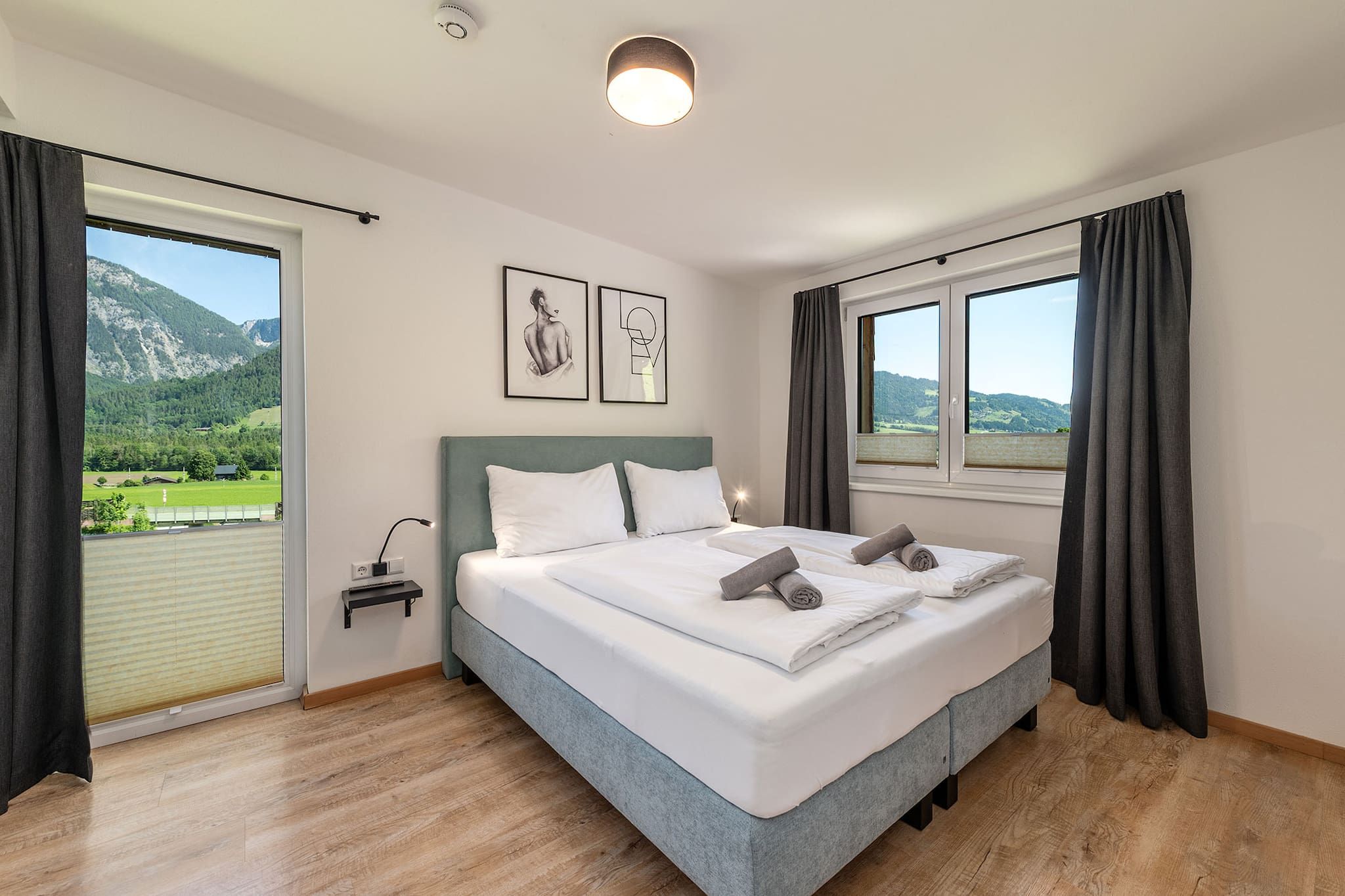 Ein Schlafzimmer mit Bett, Nachttisch, Lampe, Fenster mit Vorhängen und Blick auf eine Berglandschaft.
