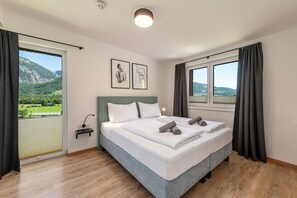 Ein Schlafzimmer mit Bett, Nachttisch, Lampe, Fenster mit Vorhängen und Blick auf eine Berglandschaft.