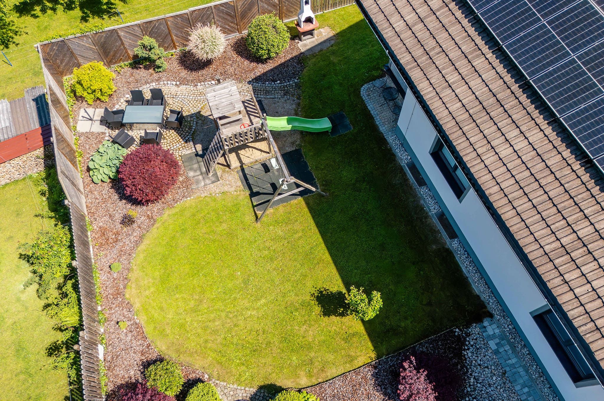 An aerial view of a residential property with a fenced garden, outdoor seating area, and a solar panel array.