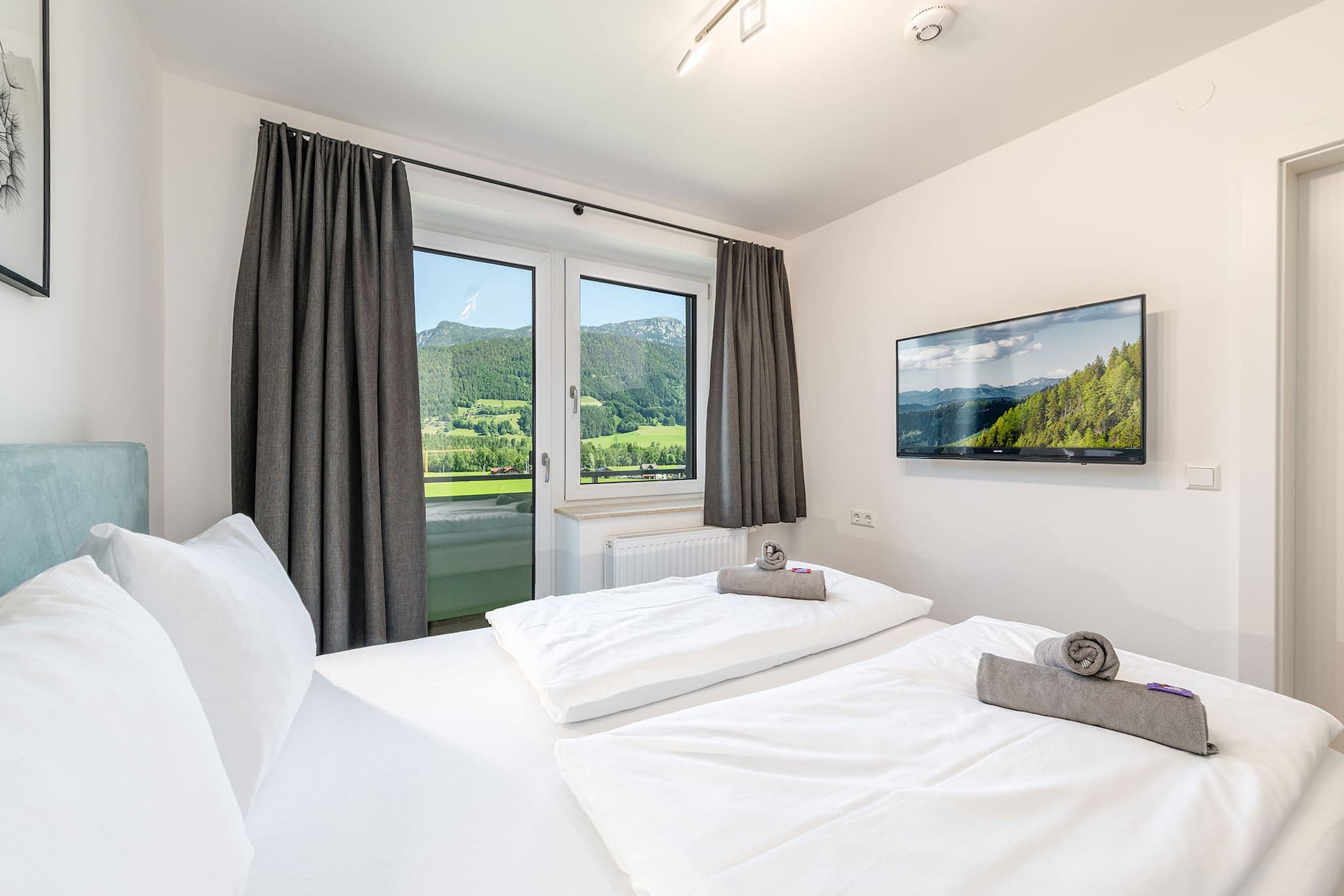 A hotel room with a bed, a window with curtains, a flat-screen TV, and a view of a mountainous landscape.