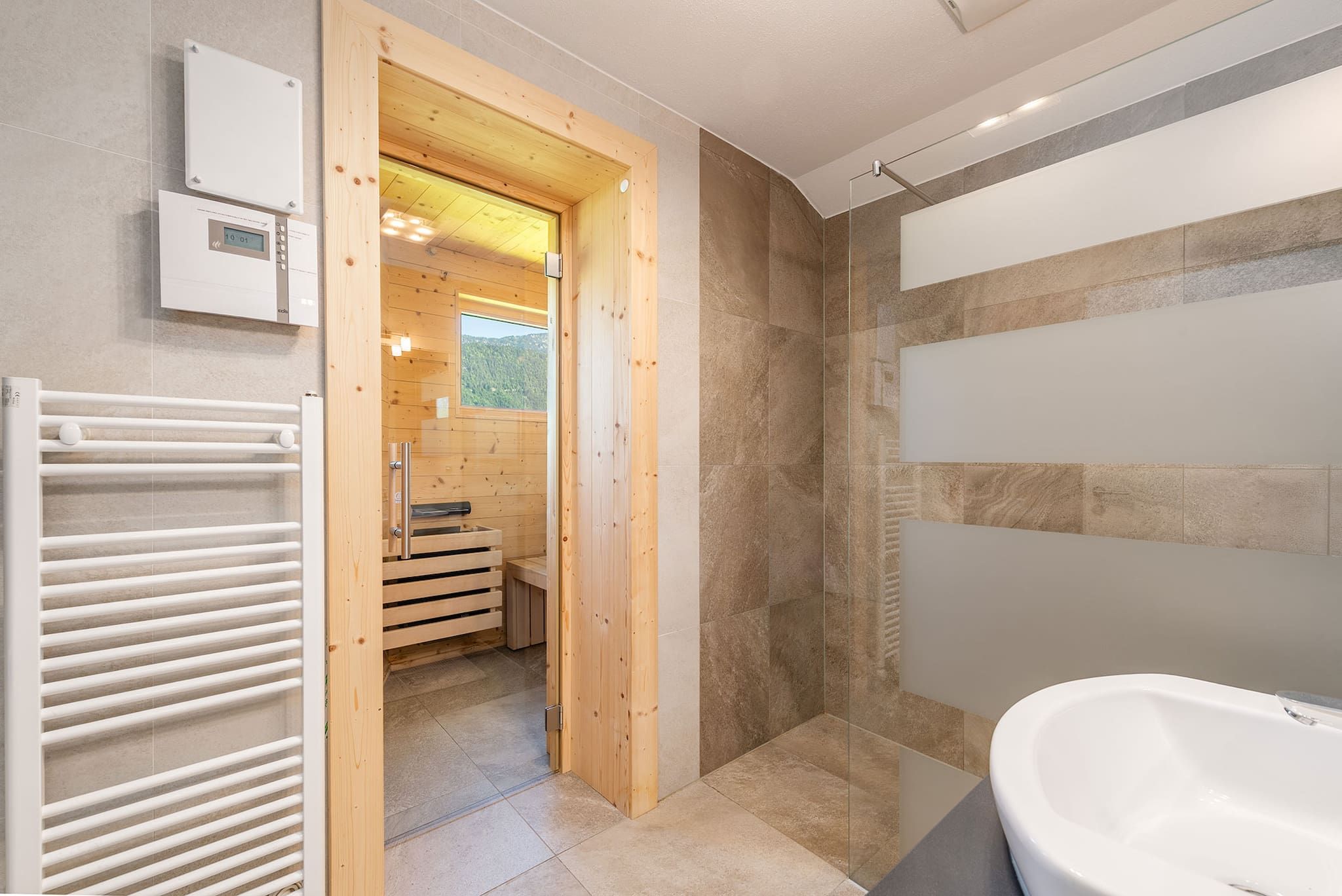Ein modernes Badezimmer mit einer großzügigen Badewanne, einer Duschkabine und einer Holztür zum Sauna-Bereich.