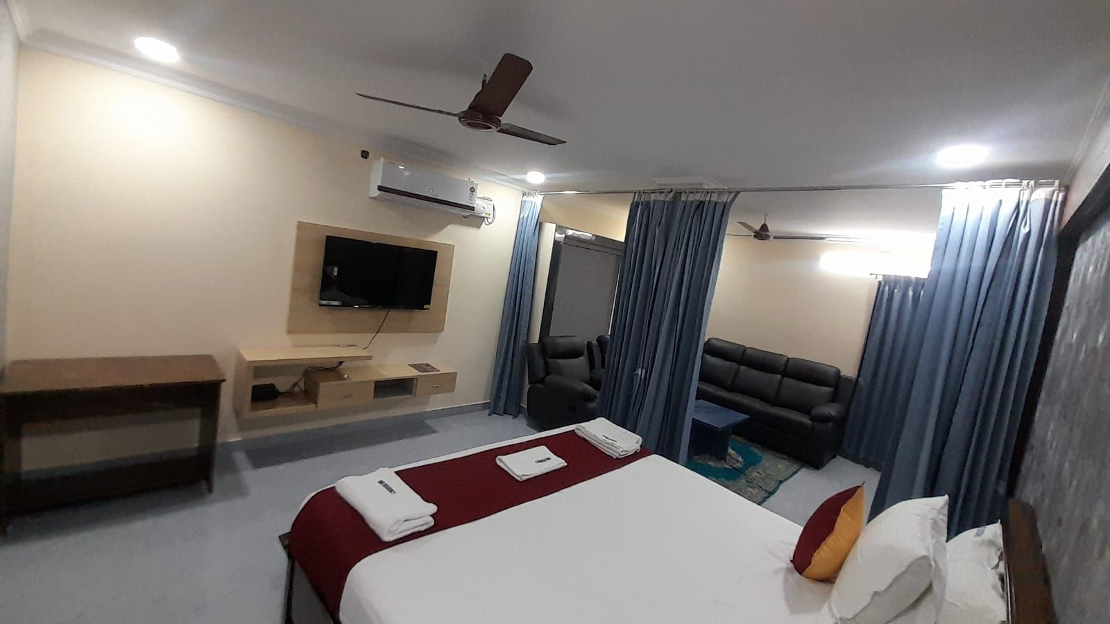 Hotellihuone, jossa on sänky, työpöytä, televisio, sohva ja kattotuuletin.