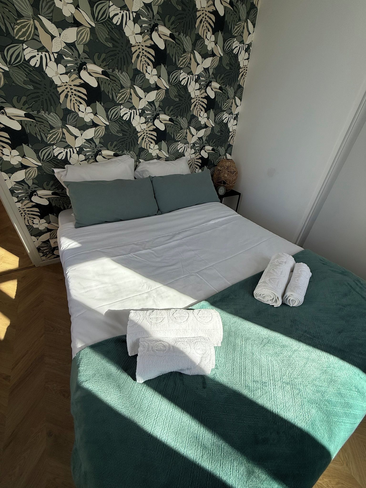 Una cama bien hecha con sábanas blancas, un plaid verde y dos toallas blancas dobladas sobre un suelo de madera.