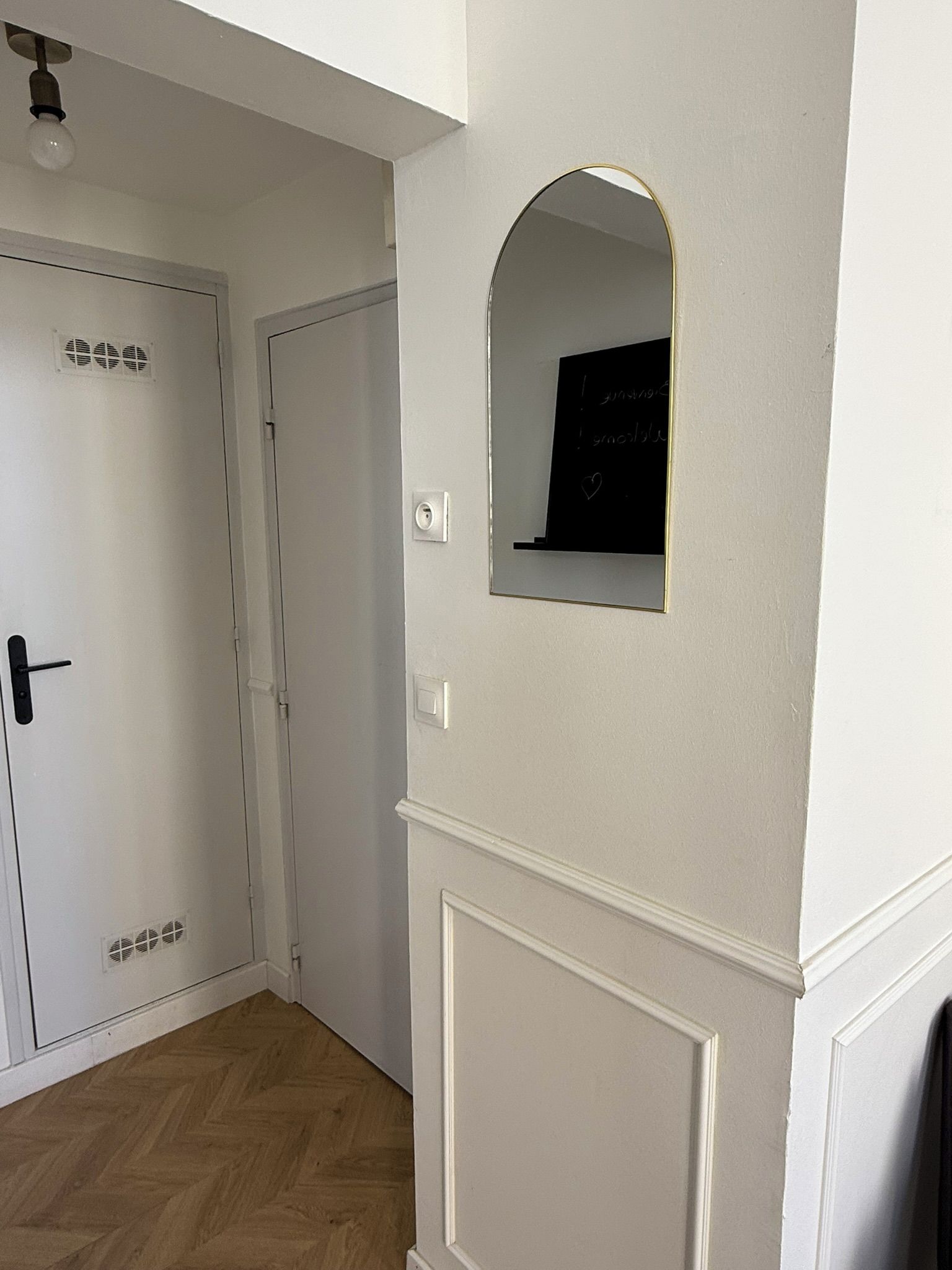 Un couloir avec une porte blanche, une petite fenêtre en arc de cercle et un luminaire fixé au mur.