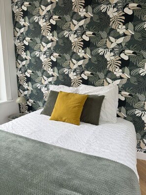 Een bed met witte lakens en een groene plaid, voor een muur met behang van tropische bladeren en vogels.