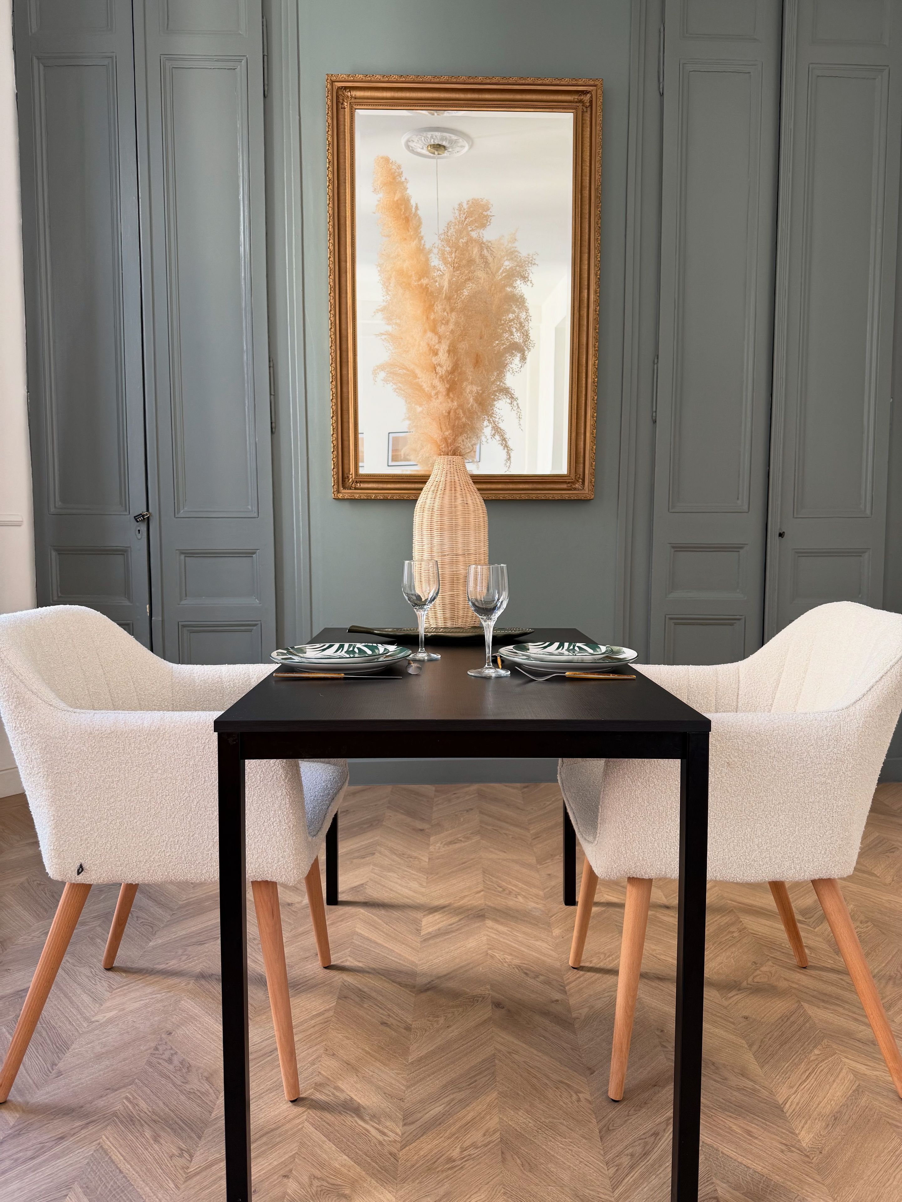 Une table à manger avec deux chaises, un vase contenant de l’herbe des pampas et un miroir encadré accroché au mur.