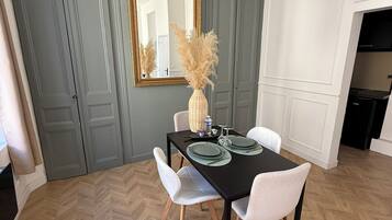 Un comedor con piso de madera en diseño espiga, una mesa negra con sillas blancas, un espejo con marco dorado y un jarrón con plantas secas.