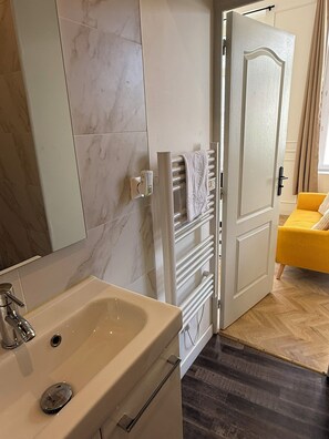 Een badkamer met een wastafel, een handdoekrek en een deur naar een kamer met een gele stoel.