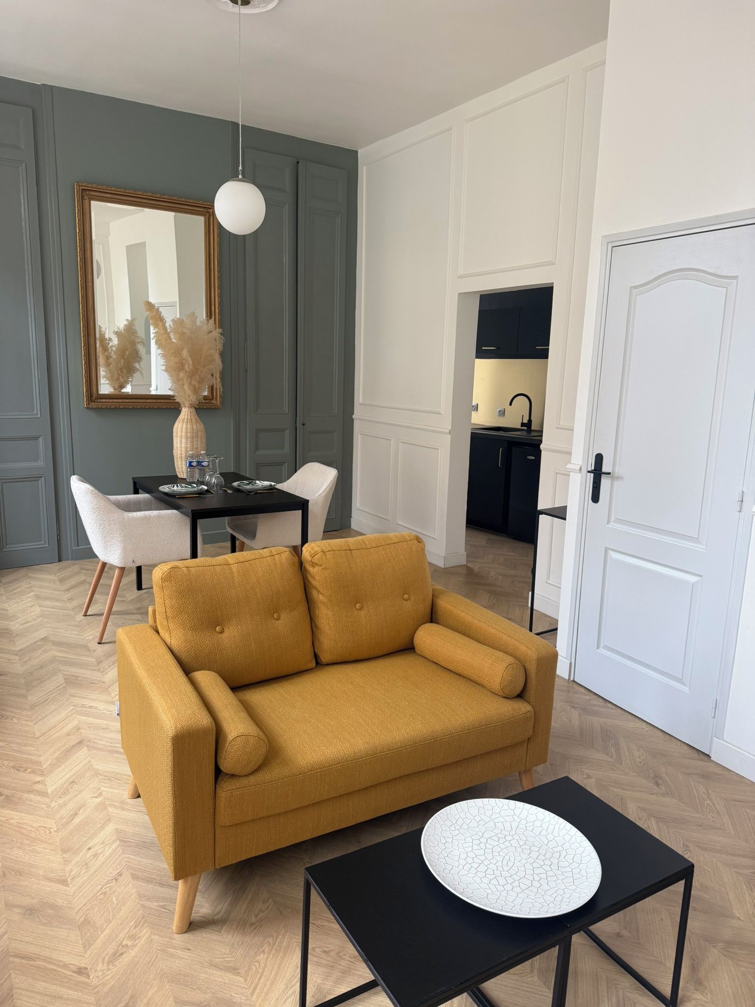 Ein Wohnzimmer mit einem senffarbenen Sofa, einem schwarzen Couchtisch, einem Esstisch mit Stühlen, einem Spiegel und einer Küche im Hintergrund.