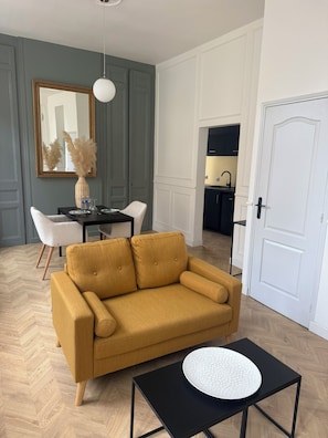 Een woonkamer met een mosterdgele bank, een zwarte salontafel, een eettafel met stoelen, een spiegel en een keuken op de achtergrond.