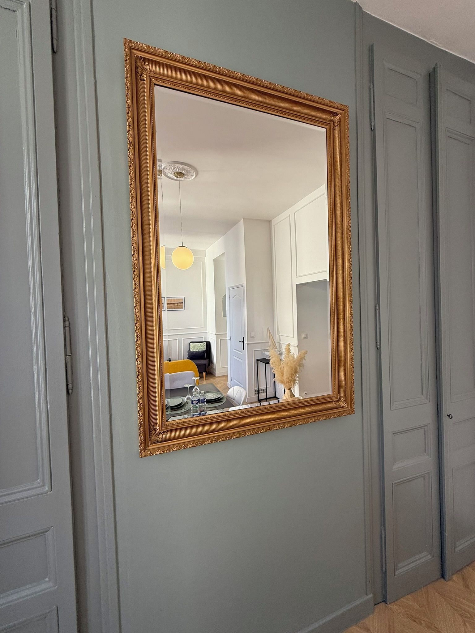 Una habitación con un espejo grande y ornamentado en la pared, una puerta, y una vista parcial de un comedor con una silla amarilla y una mesa.